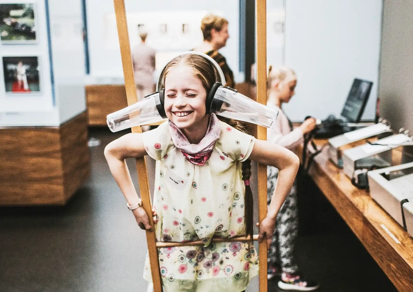 Ein kleines Mädchen steht auf Holzstelzen und hat 2 gebastelte Kopfhörer auf. Sie steht dabei strahlend und lachend im Museum, während des Stammzellspenderempfang 2019.